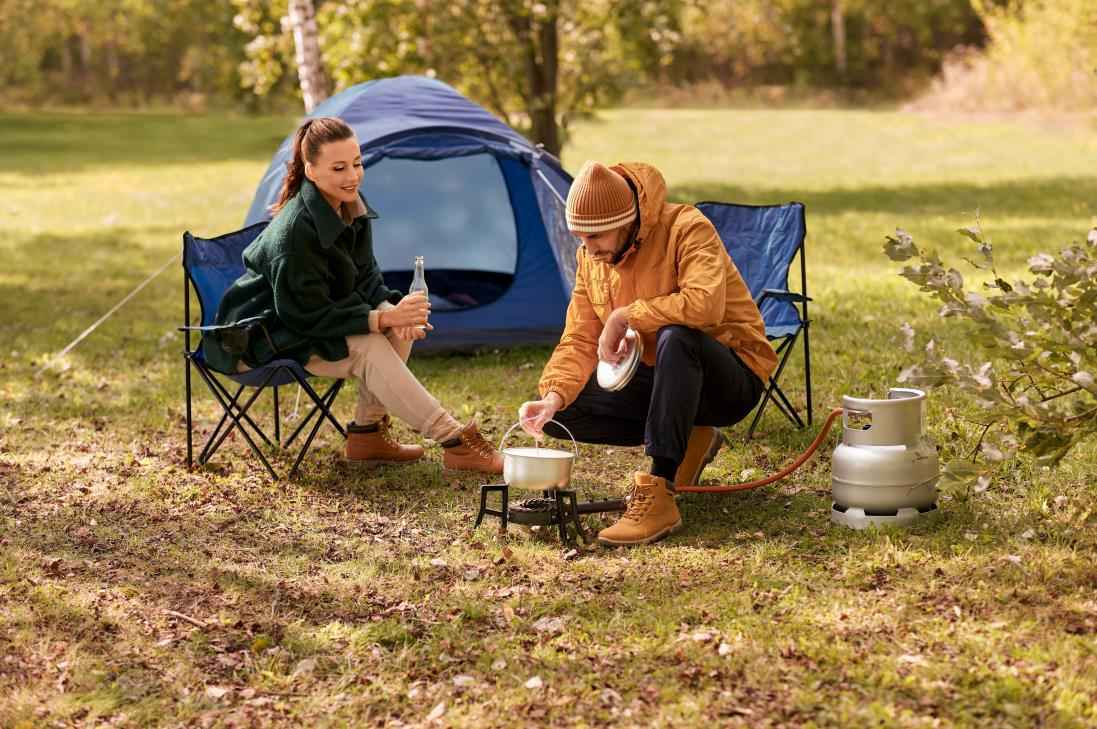 camping, tourism and travel concept - happy couple drinking beer and cooking food in pot on tourist gas burner at tent camp